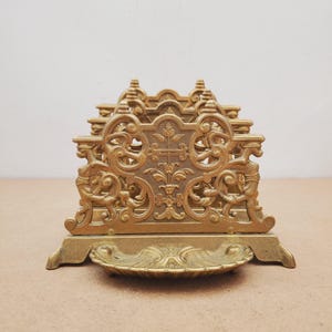 Op de afbeelding: Een goudkleurige metalen servettenhouder met een ingewikkeld, sierlijk ontwerp met krulwerk, bloemmotieven en zijfiguren. De basis bevat een klein, schelpvormig bakje aan de voorzijde, ondersteund door vier kleine pootjes.