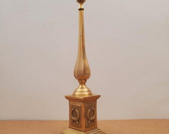 Puede incluir: Obelisco decorativo dorado. Presenta una base cuadrada con detalles de coronas de laurel, un cuerpo cónico y una parte superior decorativa. El objeto es de metal y se alza verticalmente.