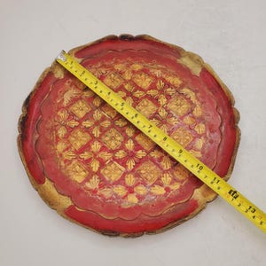 K&ouml;nnte beinhalten: Ein rundes, rotes Holztablett mit einem goldenen Design. Das Tablett hat einen gewellten Rand und ist mit einem sich wiederholenden Muster aus Blumen und Diamanten verziert.