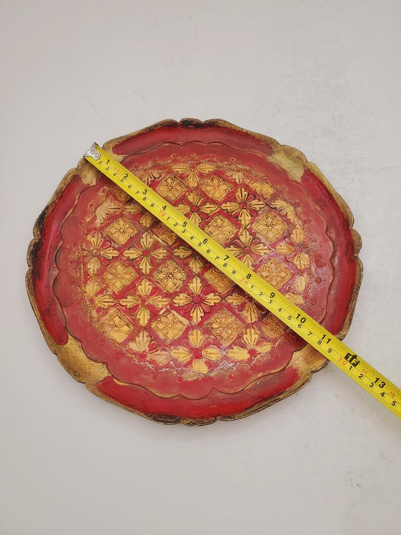 K&ouml;nnte beinhalten: Ein rundes, rotes Holztablett mit einem goldenen Design. Das Tablett hat einen gewellten Rand und ist mit einem sich wiederholenden Muster aus Blumen und Diamanten verziert.