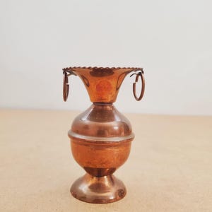 Puede incluir: Un portavelas de cobre vintage con base redonda, sección media bulbosa y parte superior acampanada con borde festoneado. Dos pequeñas asas circulares están unidas a la parte superior. El portavelas tiene un color marrón rojizo cálido.