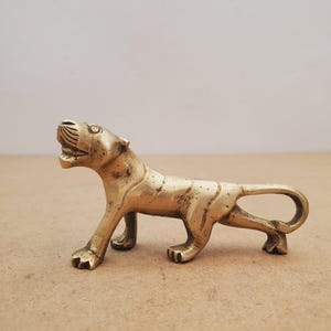 Puede incluir: Una figura de tigre de latón en posición de caminar. El tigre tiene rayas detalladas y la boca ligeramente abierta. La figura está sobre una superficie marrón claro.