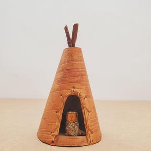 Può includere: Un piccolo teepee in ceramica marrone con una figura all'interno. Il teepee ha una forma conica con linee orizzontali e un bordo intrecciato attorno all'ingresso. Due piccoli bastoncini formano una V in cima.