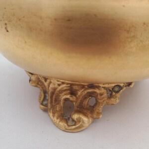May include: A close-up of a gold-coloured bowl with ornate, decorative feet. The bowl has a rounded shape and a smooth surface. The feet have a detailed, scroll-like design.