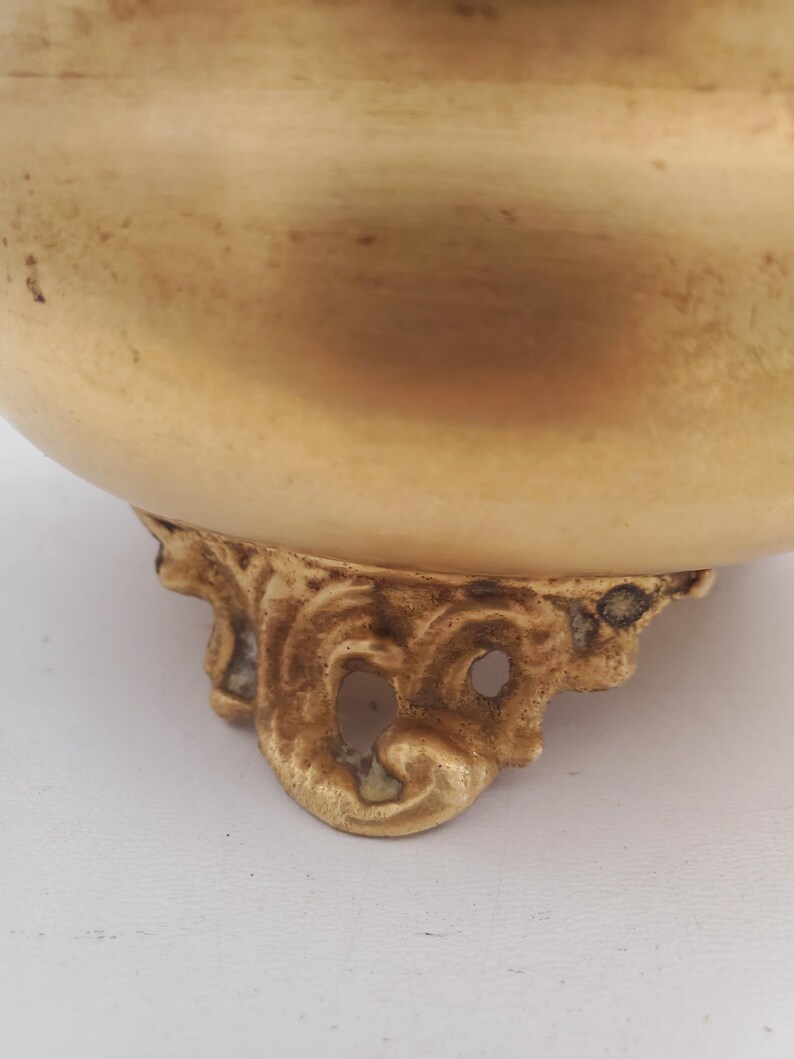 May include: A close-up of a gold-coloured bowl with ornate, decorative feet. The bowl has a rounded shape and a smooth surface. The feet have a detailed, scroll-like design.
