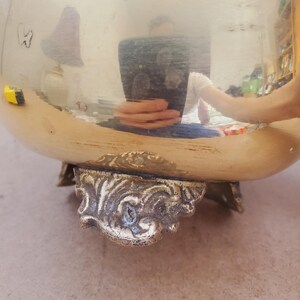 May include: A large, shiny, gold-coloured metal bowl with ornate, decorative feet. The bowl's surface reflects the surrounding environment. The feet have a detailed, antique design, adding to the bowl's vintage aesthetic.