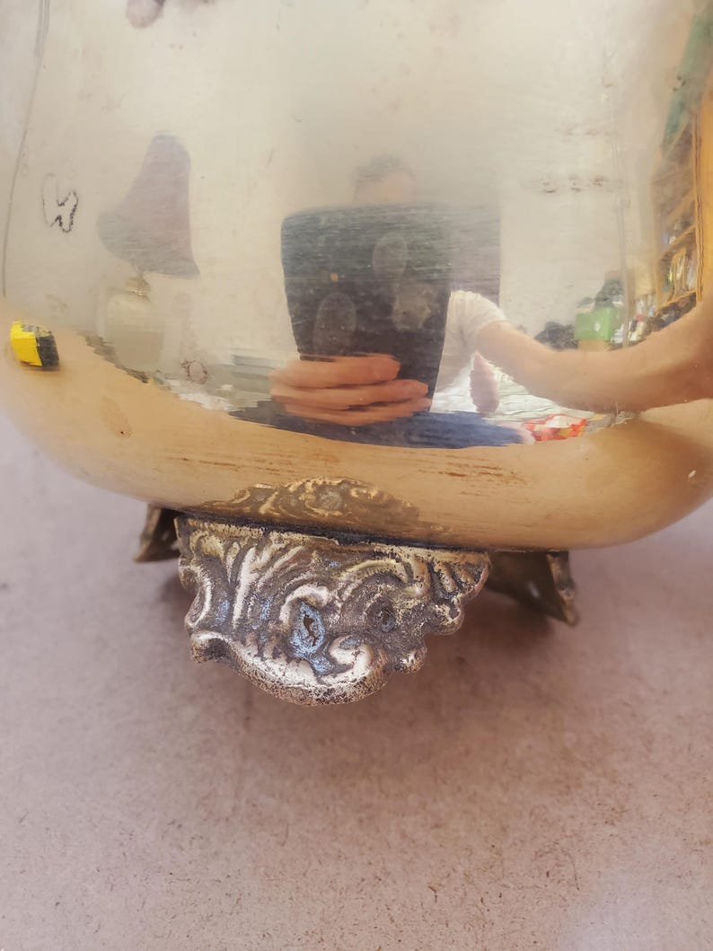 May include: A large, shiny, gold-coloured metal bowl with ornate, decorative feet. The bowl's surface reflects the surrounding environment. The feet have a detailed, antique design, adding to the bowl's vintage aesthetic.