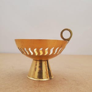 Peut inclure: Un petit bol en métal doré avec une poignée. Le bol a une base en piédestal et un motif décoratif de découpes incurvées autour du bord. La poignée est de forme annulaire simple.