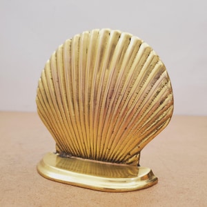 May include: A golden brass seashell-shaped decorative object. The object has a fan-like design with vertical ridges and a rounded base. The seashell is a warm, metallic color, and the base is oval-shaped.
