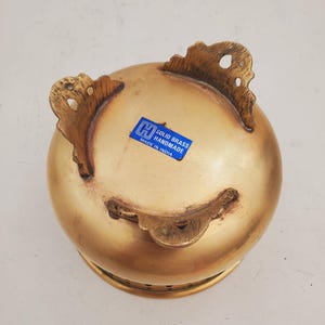 May include: A handmade, solid brass bowl with a rounded form and three decorative feet. The bowl has a golden finish and a blue sticker that reads "SOLID BRASS HANDMADE MADE IN INDIA".