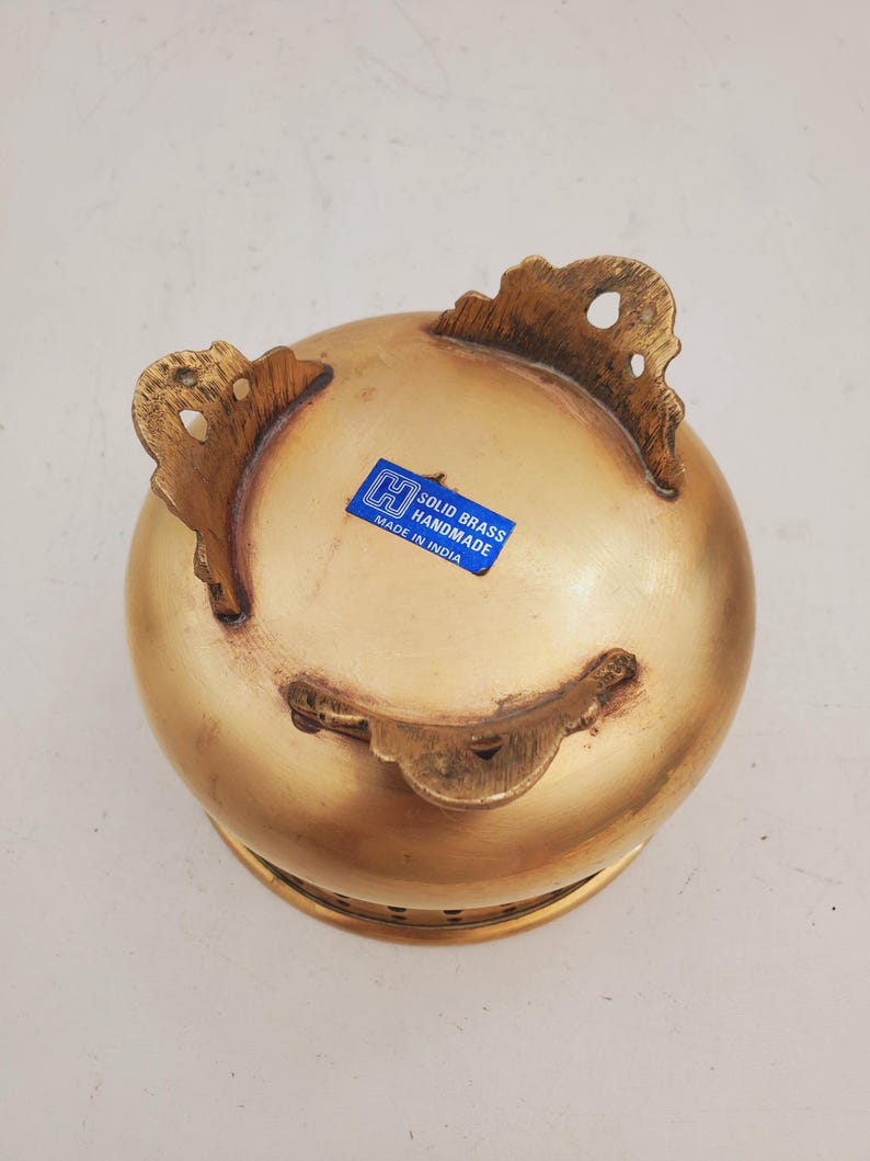 May include: A handmade, solid brass bowl with a rounded form and three decorative feet. The bowl has a golden finish and a blue sticker that reads "SOLID BRASS HANDMADE MADE IN INDIA".