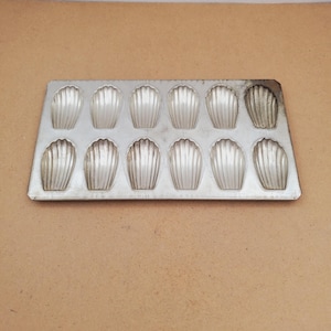 May include: A rectangular silver-colored metal baking pan featuring twelve individual shell-shaped indentations arranged in two rows of six. The surface of the pan has a metallic finish with visible signs of use and slight discoloration on one corner.