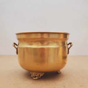 Puede incluir: Una maceta de latón pulido con un cuerpo liso y redondeado y una abertura ancha. El recipiente cuenta con dos pequeñas asas laterales ornamentadas y se apoya sobre tres pies decorativos con volutas, mostrando un acabado dorado metálico.