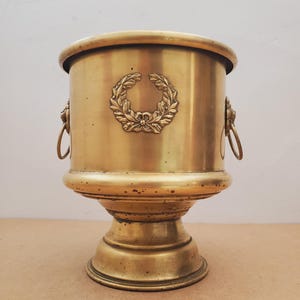 May include: A vintage brass planter with a laurel wreath emblem. The planter has a cylindrical body, a pedestal base, and two lion head handles with rings. The brass has a warm, aged patina, suggesting it is an antique.