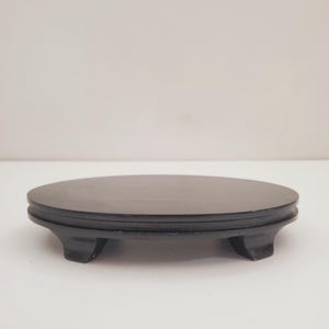 Puede incluir: Un soporte ovalado de madera negra con tres patas. El soporte está hecho de madera y tiene un acabado liso y pulido.