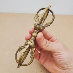 Peut inclure: Un objet rituel vajra en laiton doté d'une poignée centrale avec des bandes décoratives et deux extrémités symétriques, chacune composée de quatre griffes incurvées se rejoignant en une pointe. La surface métallique présente une patine vieillie.