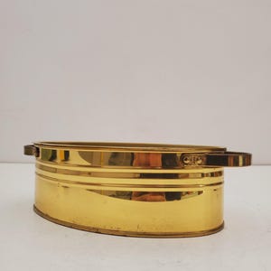 Può includere: Un contenitore ovale in ottone lucido con manici. Il contenitore ha una superficie liscia e riflettente e un design semplice. L'ottone ha una calda tonalità dorata. Misura circa 25 cm di lunghezza.