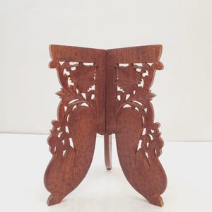 May include: A brown wooden book stand with detailed floral carvings. The stand is open, with two panels connected by a hinge. The panels feature cut-out designs and are supported by a single leg.