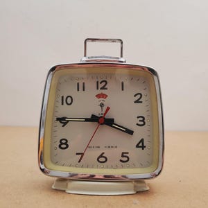 May include: A vintage square alarm clock with a cream-colored face, black hands, and red second hand. The clock has a chrome frame and a handle on top. The clock is approximately 4 inches tall.