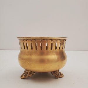 May include: A vintage brass bowl with a rounded body and decorative feet. The bowl has vertical slots around the top. The bowl is a warm gold colour.