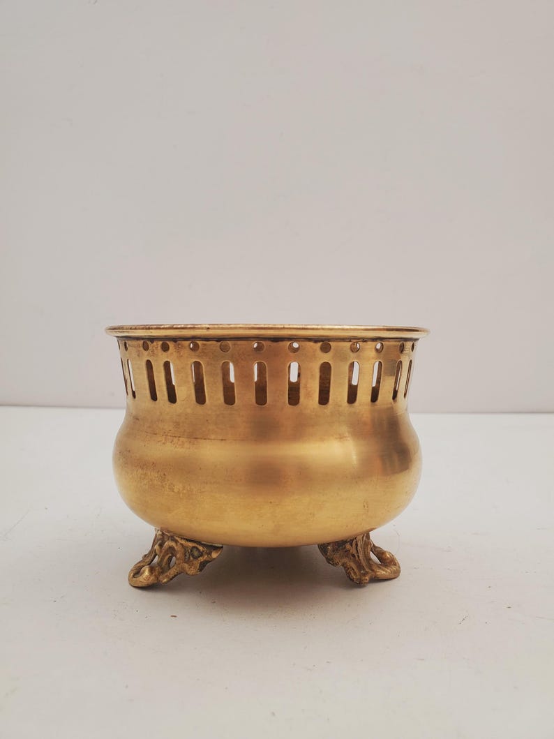 May include: A vintage brass bowl with a rounded body and decorative feet. The bowl has vertical slots around the top. The bowl is a warm gold colour.