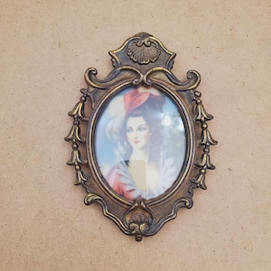 May include: An antique brass-coloured oval picture frame. The frame surrounds a portrait of a woman in a red hat and dress. The frame has decorative flourishes and a shell-like design at the top.