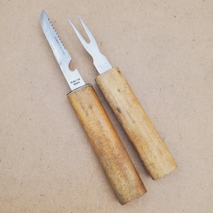 May include: Two vintage kitchen utensils: a serrated knife and a two-pronged fork. Both have wooden handles and stainless steel blades. The knife is labeled "Floating Knife" and "Stainless Steel Japan". The fork and knife are on a light brown surface.