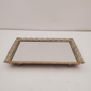 Peut inclure: Un plateau de vanité rectangulaire avec une surface miroir et un cadre filigrané doré orné. Le plateau a de petits pieds et un motif floral décoratif sur les bords. Le plateau mesure environ 30 cm de long et 20 cm de large.