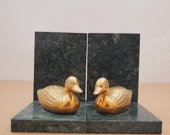 May include: A pair of bookends featuring brass duck figurines on green marble bases. The ducks are detailed with etched feathers and sit in front of dark green marble slabs. The bookends are designed to hold books upright.