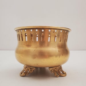 May include: A small, antique brass bowl with a rounded body and three ornate feet. The bowl has a perforated rim and a warm, golden hue. This decorative piece is likely used for display or storage.