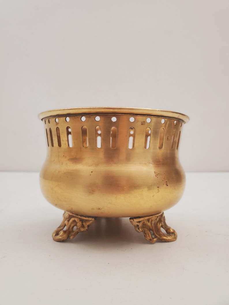 May include: A small, antique brass bowl with a rounded body and three ornate feet. The bowl has a perforated rim and a warm, golden hue. This decorative piece is likely used for display or storage.