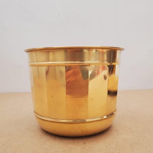 Op de afbeelding: Een cilindrische, goudkleurige metalen plantenbak met een licht afgeronde basis en een gerolde rand. De plantenbak heeft verticale ribbels en een gepolijste afwerking, die het licht reflecteert. Geschikt voor binnen of buiten.
