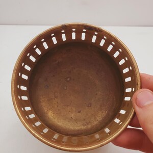 May include: A small, circular brass bowl with a pierced edge. The bowl has a smooth, aged appearance and is held by a hand. The interior of the bowl is a darker shade of brass. The bowl is likely a decorative or functional item.
