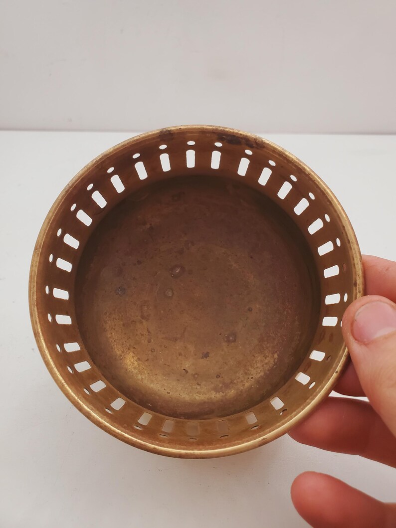 May include: A small, circular brass bowl with a pierced edge. The bowl has a smooth, aged appearance and is held by a hand. The interior of the bowl is a darker shade of brass. The bowl is likely a decorative or functional item.