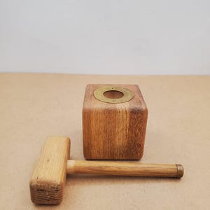 Puede incluir: Un mazo de madera y un bloque de madera cuadrado con un anillo de latón incrustado en la parte superior. El mazo tiene una cabeza rectangular y un mango cilíndrico. El bloque y el mazo son de madera clara.