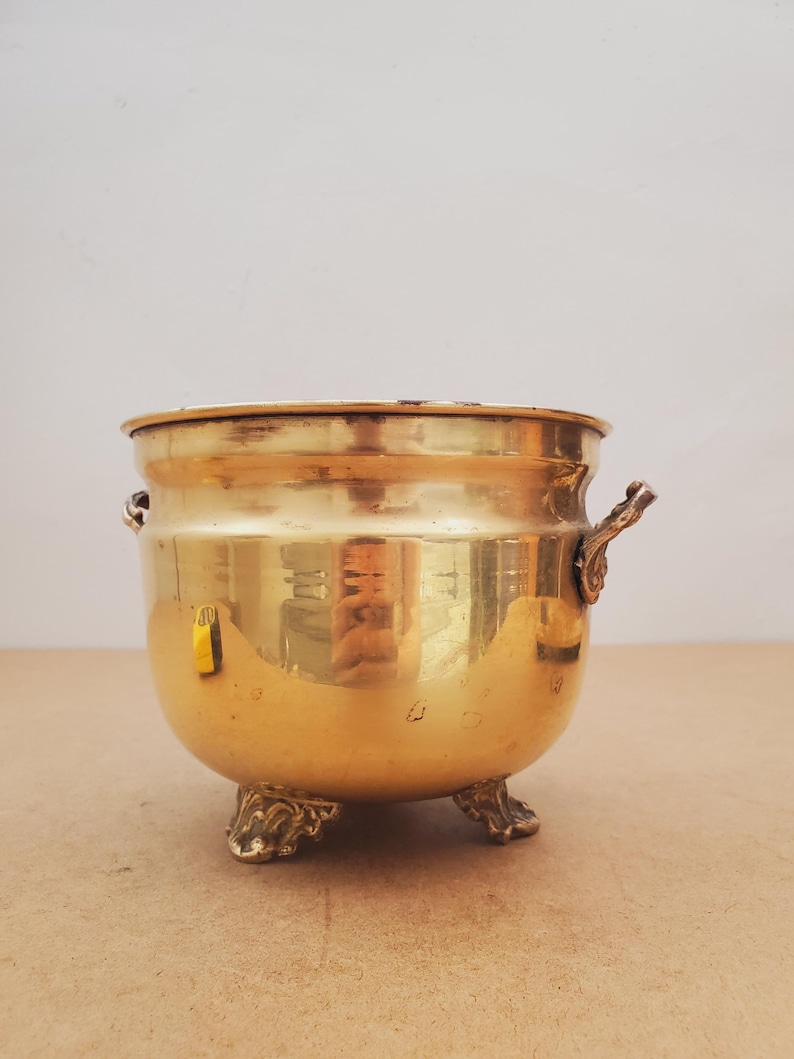 May include: A polished brass cauldron, golden in colour, with decorative handles and feet. The round pot has a wide opening and a curved body. The surface reflects the surroundings, creating a warm aesthetic. The cauldron is likely used for decorative purposes.