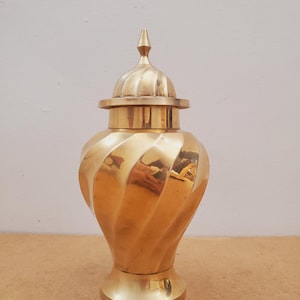 Może przedstawiać: Złoty, dekoracyjny wazon z pokrywką. Wazon ma spiralny wzór i spiczasty szczyt na pokrywce. Metalowy wazon stoi na płaskiej powierzchni.