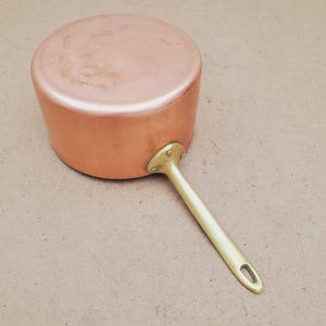 May include: A round copper saucepan with a smooth, polished finish and a flat base. The pot features a long, sturdy brass handle attached to the side with two visible rivets. The handle includes a small, oval-shaped hole at the end for hanging.