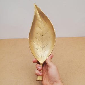 May include: A gold-coloured leaf-shaped decorative tray. The tray has detailed veining and a stem-like handle. The leaf is held by a person's hand against a neutral background. The tray is made of metal.