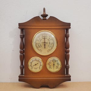 Puede incluir: Una estación meteorológica de madera con un barómetro y dos medidores. El barómetro tiene una esfera circular con una aguja y texto que indica las condiciones meteorológicas. Los medidores muestran lecturas de temperatura y humedad. El marco es de color marrón oscuro.