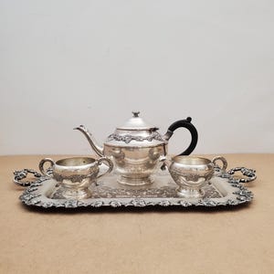 May include: A silver tea set on a decorative tray. The set includes a teapot with a black handle, a creamer, and a sugar bowl. The tray and tea set have ornate floral detailing.