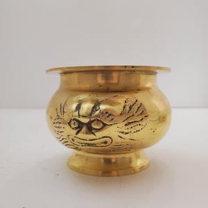 Puede incluir: Un cuenco pequeño y redondo de color latón con un borde acampanado y una base de pedestal. El cuenco presenta un diseño grabado de una cara estilizada. El cuenco probablemente se utiliza con fines decorativos o como un recipiente pequeño.