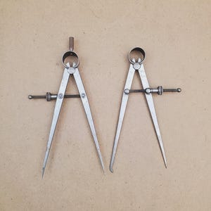 May include: Two vintage metal calipers with pointed tips, used for measuring distances. The calipers have a silver finish and are set against a neutral background. Each caliper has an adjustment screw and a curved top.