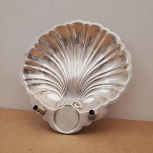 May include: A silver-colored, shell-shaped decorative tray. The tray has a scalloped edge and a central, circular indentation. Two small spouts and a circular base are visible. The tray is made of a shiny, reflective metal.