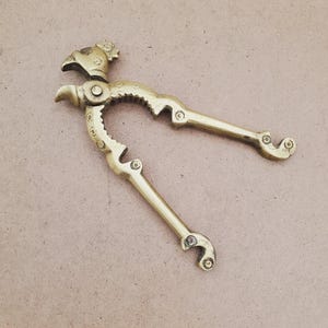 May include: A vintage brass nutcracker with a bird-like design. The nutcracker features a detailed bird head at the top and curved handles. The brass has a warm, golden hue, and the tool is designed to crack nuts.