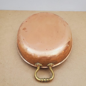 Puede incluir: Una sartén ovalada de color cobre con un asa de latón. La sartén tiene un aspecto ligeramente desgastado, con algunas manchas más oscuras en la superficie. El asa tiene un diseño curvo y ornamentado. Esta batería de cocina vintage es ideal para cocinar.