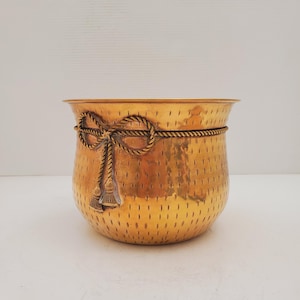 Puede incluir: Un macetero de metal dorado con un diseño de cuerda alrededor de la parte superior y borlas en los lados. El macetero tiene una textura martillada.
