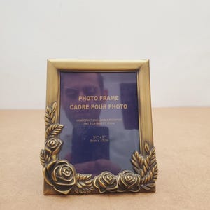 May include: A rectangular, antique gold-colored photo frame. The frame is decorated with rose and leaf motifs. The text "PHOTO FRAME CADRE POUR PHOTO" is visible. The frame is 3.5" x 5" (9cm x 13cm).