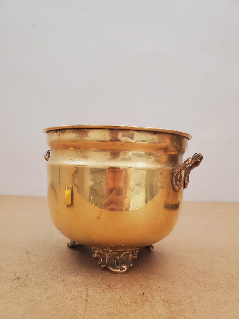 May include: A polished brass planter, featuring a rounded body and decorative feet. The planter has two ornate handles and a wide opening. The brass reflects light, creating a warm glow. This vintage piece is ideal for displaying plants or as a decorative accent in a home.
