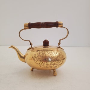 Peut inclure: Une théière dorée avec un motif floral et une poignée en bois. La théière a trois pieds et un petit bouton sur le couvercle.